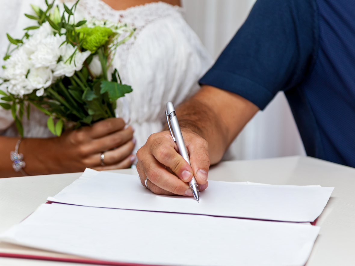Hochzeit: Die Braut hält einen Blumenstrauß, der Bräutigam unterschreibt auf einem Dokument.